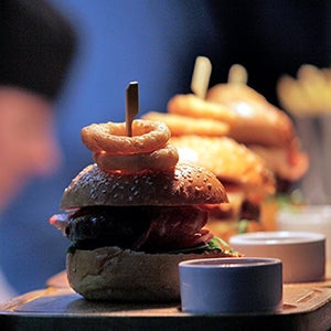 A Clydebuilt burger with onion rings on top on a wooden board with a ranekan of sauce on the side.