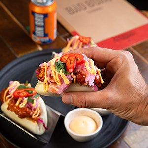 A person is holding a bao bun with various fillings inside, above a plate with another bao bun and condiment. Next to the plate is a can of Irn-Bru and a menu.