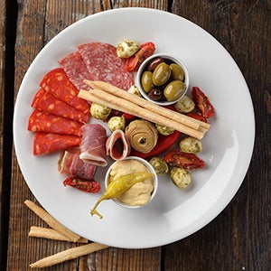 A charcuterie plate including; cured meats, olives, sundried tomatoes, mozzarella, bread sticks and a condiment.