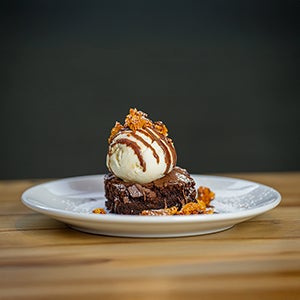 A plate with a slice of brownie and a scoop of ice cream on top with sauce and biscuit crumbled over it.