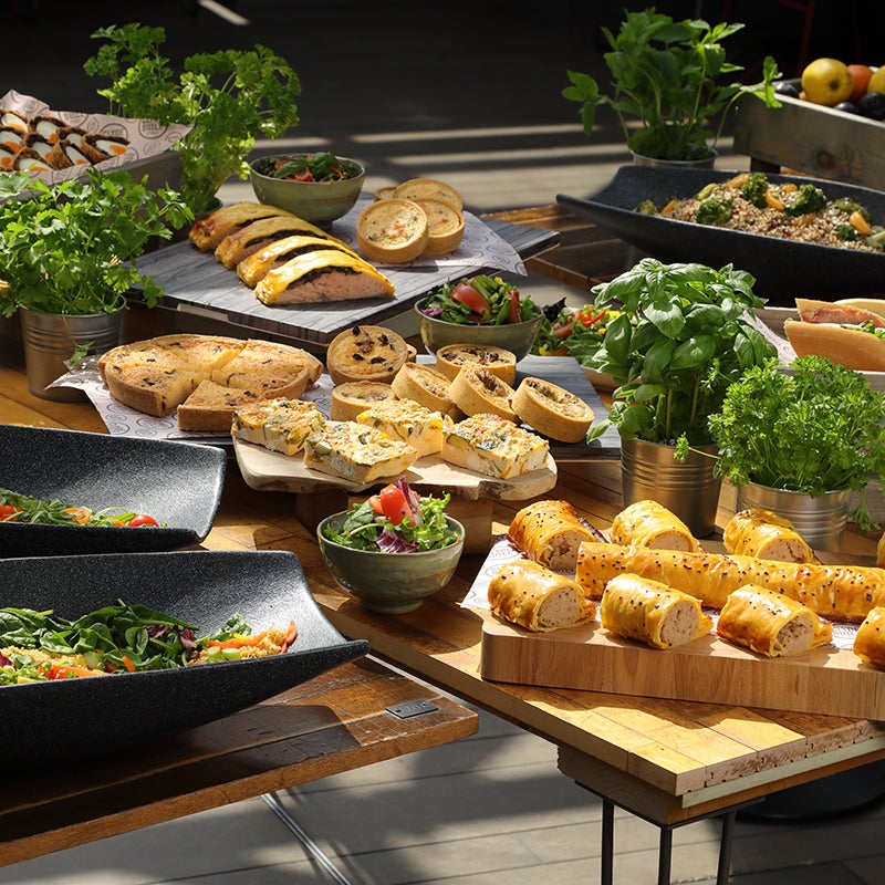 A farmer market spread of food on a wooden table. This consists of various foods like salads, sausage rolls, quiche and sandwiches.