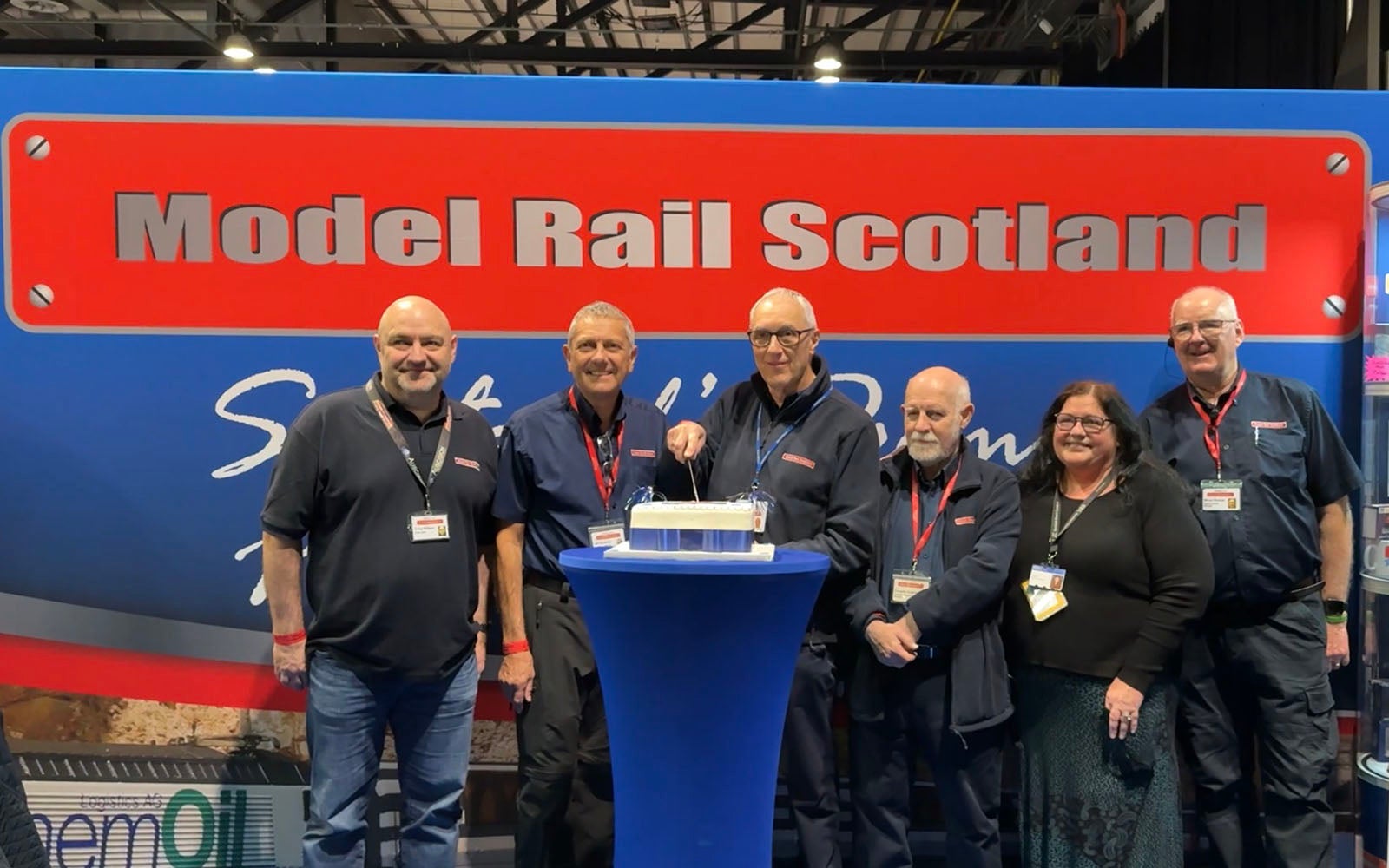 A group of people standing in front of Model Rail Scotland Signage cutting a cake.