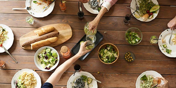 Birdseye view of numerous plates of food and drinks on a table. People taking drinks and food can be seen.