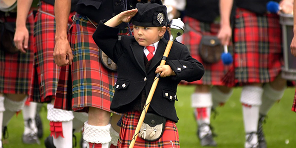 Child wearing traditional Scottish clothing including a kilt, saluting with an instrument in hand.