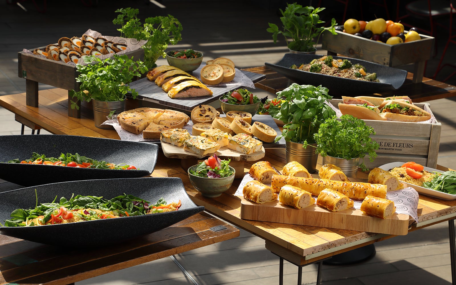 A farmer market spread of food on a wooden table. This consists of various foods like salads, sausage rolls, quiche and sandwiches.